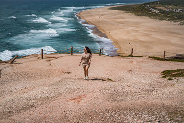 A girl is walking, admiring the calm ocean in Nazare, beachside in the summertime, Portugal