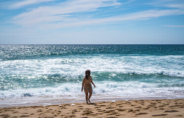 A girl is walking and looking back next to the calm ocean in Nazare, summertime, Portugal