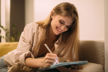 Work at home. A young blonde girl draws on a tablet while sitting on the sofa.