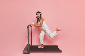 Lady in white silk pajamas and pink sleepers on treadmill against pink background, holding mug in hand, lifestyle concept, copy space