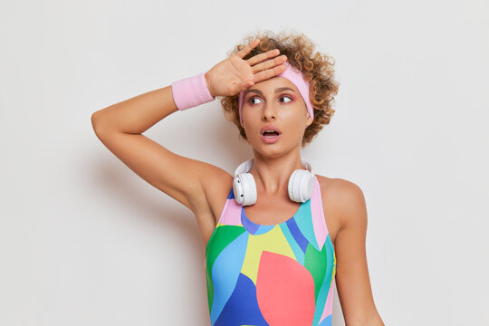 Curly sportswoman in colored bodysuit against white background, headphones on her neck, having rest after workout, sports fashion concept, copy space
