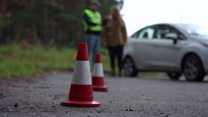 Close-up red and white road cones with blurred male instructor teaching female student standing at car at background. Unrecognizable Caucasian man talking with woman explaining driving lesson