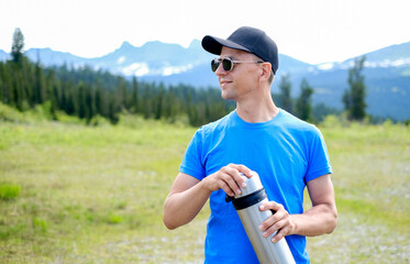 A male tourist with thermos in mountain background. A tourist drinks coffee from a thermos in the...