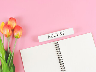 flat lay of opened diary or notebook with wooden calendar August on pink background with orange-yellow tulips.
