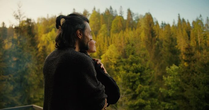 A brunette man approaches a girl on the balcony of a country house, hugs her and together they look at the mountains covered with coniferous forest