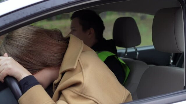 Stressed sad young woman leaning on steering wheel crying as man talking sitting at background in car. Desperate female Caucasian student failing driving exam