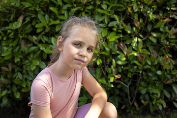 Portrait of a cute and pretty blonde little girl against a background of green broad-leaved plants....