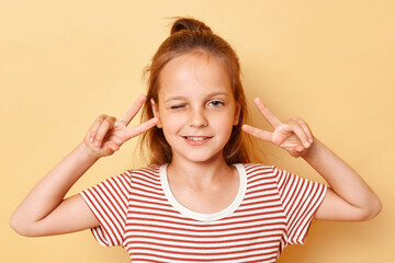 Dreamy cheerful dark haired little girl showing v-sign near eyes winking wearing casual style striped T-shirt standing isolated over beige background.