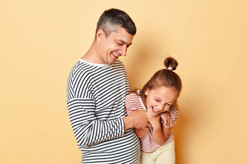 Father and daughter having fun together isolated over beige background family wearing striped shirts dad tickles little kid and laughing happily.