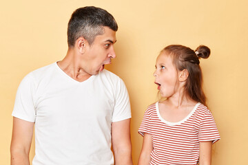 Surprised amazed astonished father and daughter wearing casual t-shirts standing isolated over beige background looking at each other with widely opened mouths.