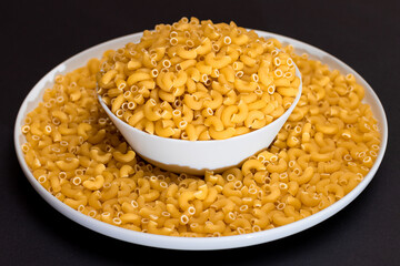 Uncooked Chifferi Rigati Pasta in White Plate on Black Background. Fat and Unhealthy Food. Scattered Classic Dry Macaroni. Italian Culture and Cuisine. Raw Pasta