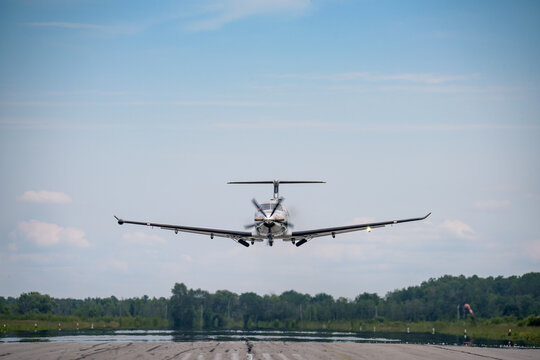 A small business turboprop Pilatus PC-12 on final with the landing gear starting to extend.  Blue sky with haze and clouds in the background.