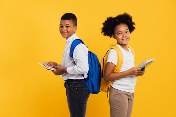 Happy african american schoolgirl and schoolboy holding books side by side on yellow background....