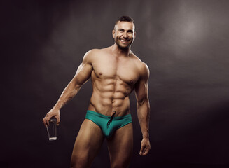 Fototapeta na wymiar Tanned sports man in turquoise swimming trunks with glass in his hands. Smiling male model posing in the studio. Sexy bodybuilder with six pack abs in underwear in studio.