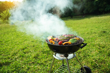 Grilled meat in grill. Roasting steaks and vegetables bbq in a round grill in a meadow. Summer picnic outdoors.