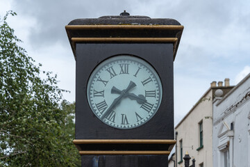 Fototapeta na wymiar Authentic vintage street clock, Roman numerals, time 3:37, black hands against a cloudy sky. Green birch branches on the left, an old building wall on right. Clock hands are in the shape of swords.