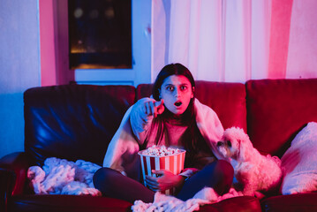 Beautiful young girl at home in a dark room with a popcorn bucket playing with her dog and sits in front of a monitor or TV.