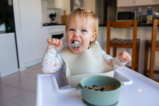 One year old baby girl using spoon to eat weetbix and sultanas for breakfast