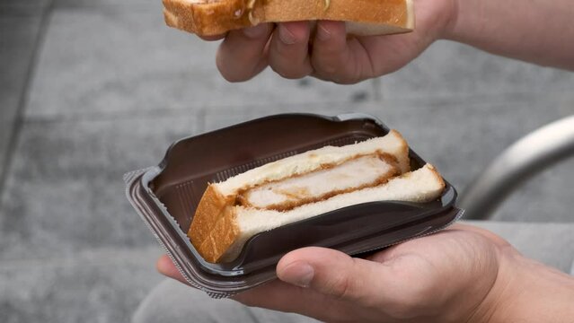 Unrecognizable person eating katsu sando, Japanese sandwich with pork cutlet, cabbage and tonkatsu sauce, at street.