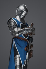 A medieval soldier garbed in blue surcoat and armor holding a sword with a still, statue-like pose,...