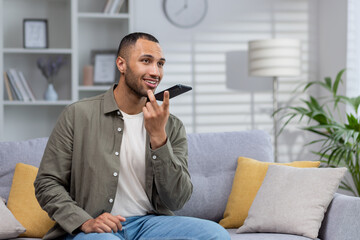 African american young man sitting on sofa at home and talking on speaker phone, recording voice message, dictating his conversation on mobile app