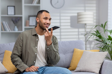 African american young man sitting on sofa at home and talking on speaker phone, recording voice...
