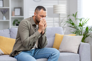 African American man sick at home. He sits on the couch and blows his nose into a tissue - Powered by Adobe