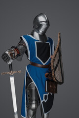 Fototapeta premium Medieval guard dressed in armor and blue surcoat holding a sword, standing in statue pose, against a gray background