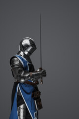 Naklejka premium A medieval soldier garbed in blue surcoat and armor holding a sword with a still, statue-like pose, against a gray background