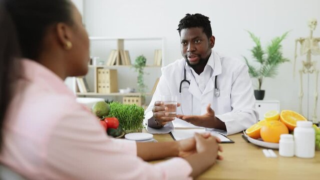 Attractive bearded man in doctor's coat gesturing at glass with fluid while meeting with multiethnic patient. Smiling nutritionist recommending clean water as help in reducing calorie intake on diet.