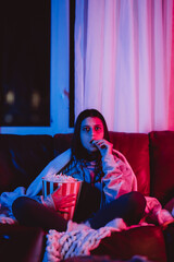 Beautiful young girl eating popcorn watching movie at home in a dark room with a popcorn bucket and playing with her dog and sits in front of a monitor or TV.