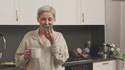Medium shot of modern senior Asian woman recording voice message on smartphone standing at modern kitchen at daytime holding cup of coffee in hand - Powered by Adobe