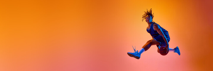 Athletic young man, basketball player in motion, jumping with ball against orange background in...