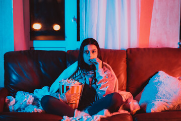 Naklejka premium Beautiful young girl eating popcorn watching movie at home in a dark room with a popcorn bucket and playing with her dog and sits in front of a monitor or TV.