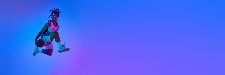 Dynamic image of young basketball player in motion, jumping with ball against gradient blue...