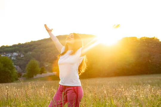 A woman stands in a field in nature with her hands raised in the rays of the setting sun in summer.
