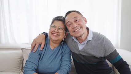 Happy elderly asian couple embracing bonding smiling and looking at camera while sitting on comfortable sofa in living room at home. Couple retirement lifestyle
