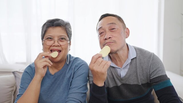 Happy Elderly Asian Couple Enjoy Eating Potato Crisps While Sitting On Comfortable Sofa In Living Room At Home. Couple Retirement Lifestyle