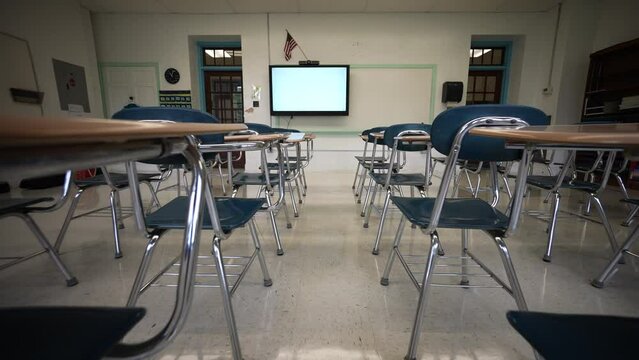 Camera pushes forward through a row of empty desks chairs with smart board, whiteboard, marker board and US flag.