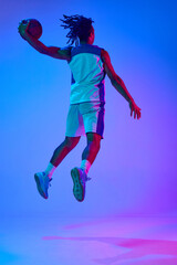 Fototapeta na wymiar Full-length dynamic image of young professional athlete, basketball player in motion during game against gradient blue background in neon. Concept of professional sport, competition, competition, ad
