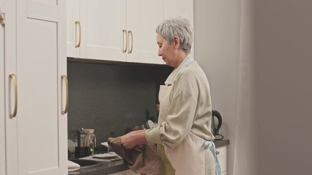 Medium shot of grey haired senior Asian woman drying dishes with towel and putting them on lowest shelf of white kitchen cabinet