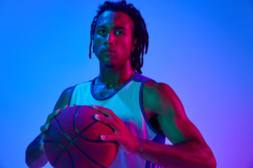 Young man with dreads, basketball player in uniforma posing with ball against gradient blue...