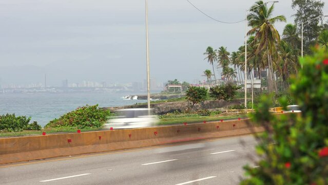 Timelapse of fast-moving cars and trucks in the foreground on the speed highway road, with downtown Santo Domingo and palm trees in the background. Busy life in megapolis