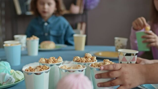 Close-up selective focus shot of marshmallow and sweets on table at birthday party, young children sitting around and eating, and hand taking glass with popcorn