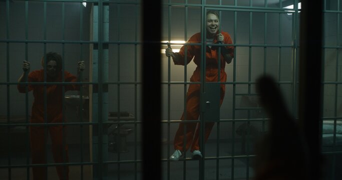 Aggressive female prisoner in orange uniform yells at neighbor inmate, holds metal bars in prison cell. Women serve imprisonment terms for crimes in jail. Detention center or correctional facility.