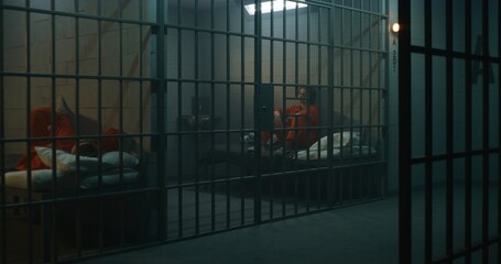 One female prisoner in orange uniform stands behind metal bars, another sits on the bed in prison...