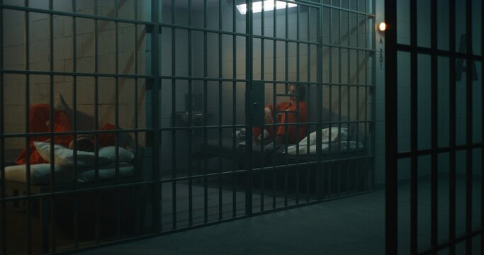 One female prisoner in orange uniform stands behind metal bars, another sits on the bed in prison cell. Women serve imprisonment terms for crimes in jail. Depressed inmates in detention center.