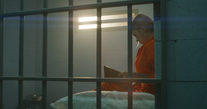 Female prisoner in orange uniform sits on bed behind metal bars, reads Bible in prison cell, looks at barred window. Woman criminal serves imprisonment term for crime in jail or correctional facility.