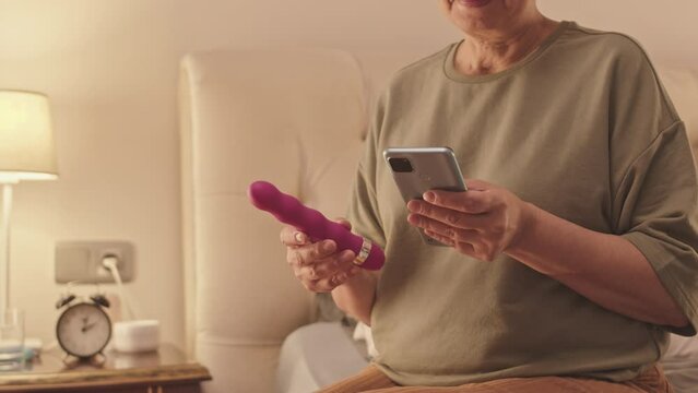 Cropped shot of senior woman sitting on bed at home alone browsing on smartphone holding purple vibrator in hand