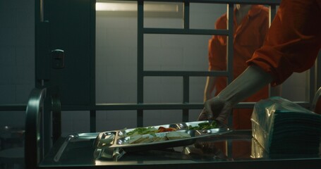 Prison worker gives food from serving trolley to female prisoner in orange uniform in jail cell. Woman criminal sits on bed, eats dinner. Imprisonment term in prison or detention center. Dolly shot.